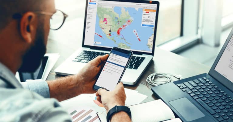 Man checks his phone to read a hurricane notification on his AlertMedia app while the AlertMedia platform displaying the path of a hurricane on his laptop