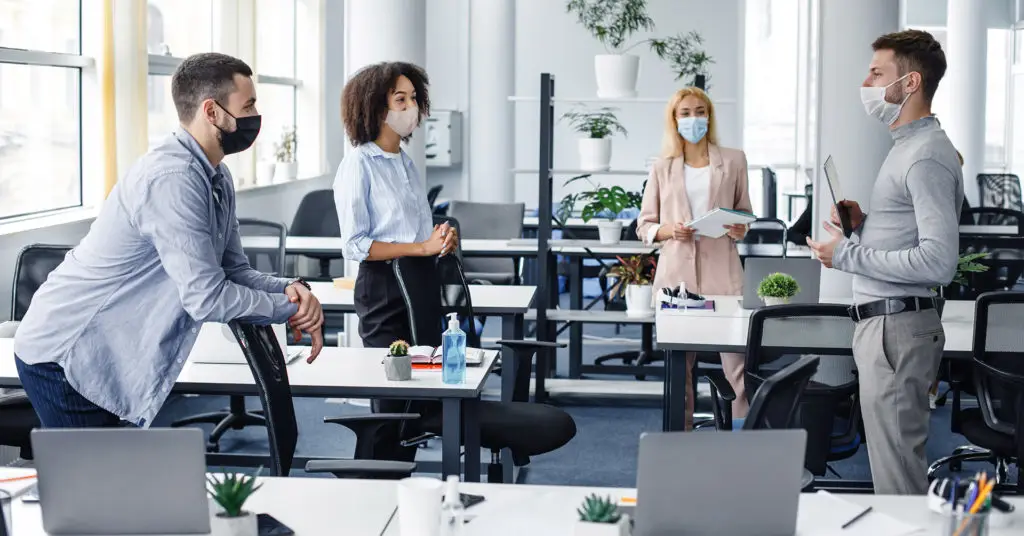 four office workers wearing masks discuss plans for 2021 while standing several feet apart