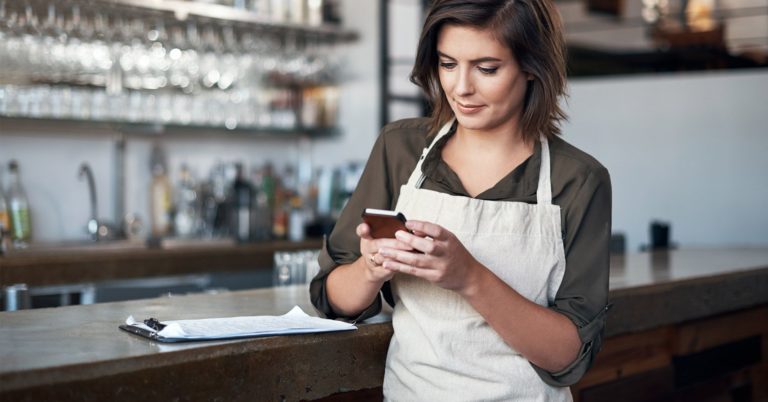 a female bar manager wearing an off-white apron looks at her phone while a clipboard sits on the counter to her right
