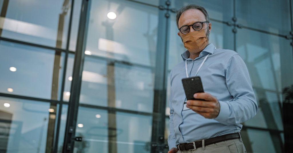 A middle-aged man wearing round, acetate glasses, an ID badge on a lanyard, and on a face mask checks a notification on his phone.