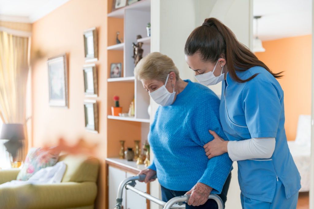 Home caregiver wearing a facemask while assisting a senior woman walking with an elderly woman.