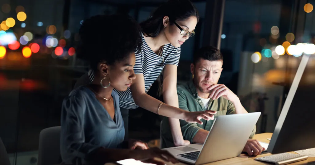 Three co-workers look at a laptop together, reviewing an emergency plan late in the office.