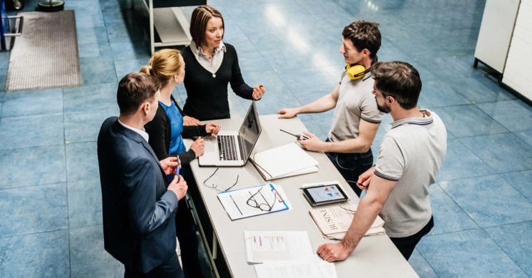 A team of 5 workers standing around a table, having a discussion