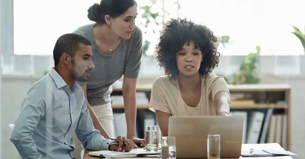 Three coworkers look at a laptop screen together, discussing urgent preparedness initiatives.