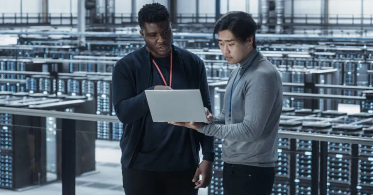 Two workers look at a laptop together, reviewing IT risk mitigation procedures.