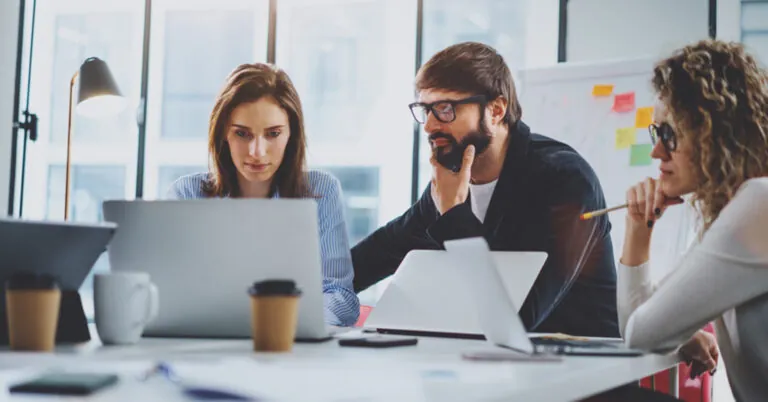 Three people gather around a conference table with laptops to discuss risk management.