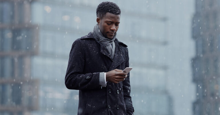 A man looks down at his phone as snow falls around him.