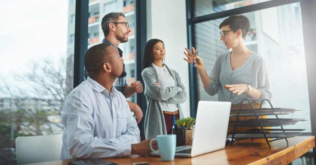 A team of co-workers gather around a desk as a woman describes plans and gestures with her hands.