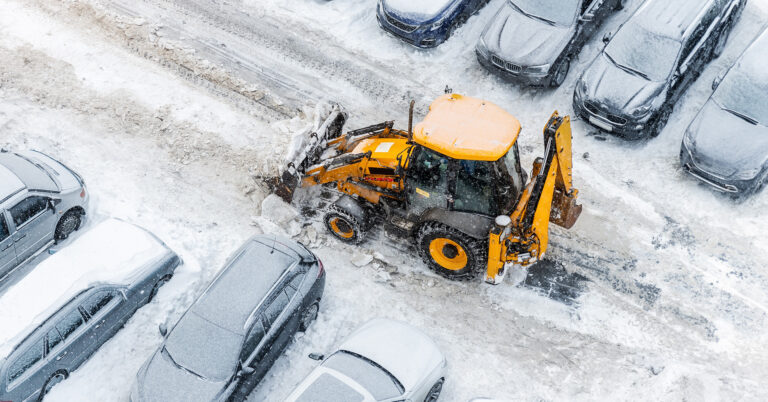 A backhoe loader tractor plows snow in a parking lot, passing between rows of parked cars.