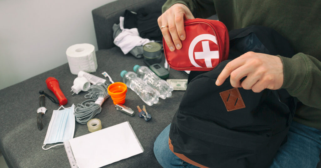 A person has emergency supplies spread out and is adding them to a backpack