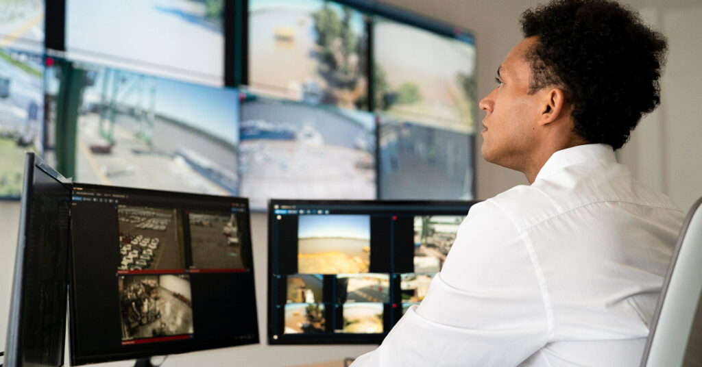 Security specialist sits at a desk watching live facility views on desktop and wall screens