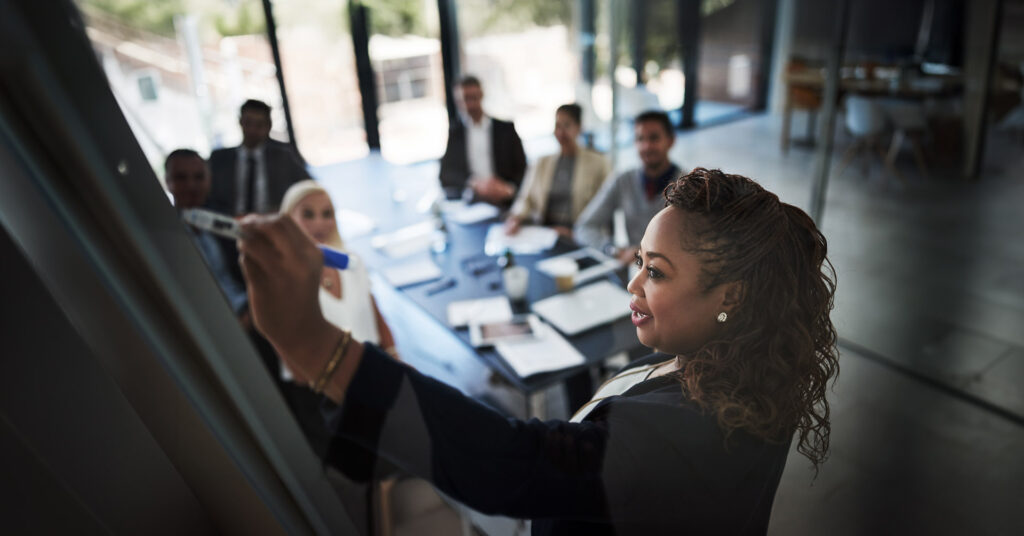 A woman uses a white board to guide a discussion of business continuity strategy with her team in a conference room