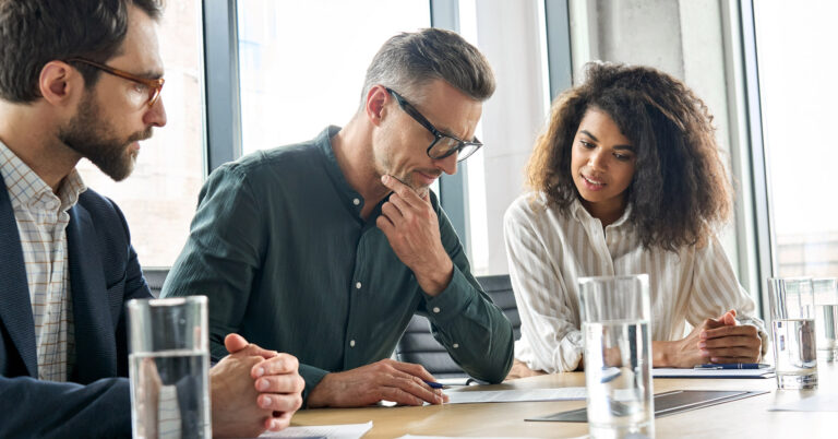 Three colleagues sit at a table, collaborating on a paper document that sits in front of one man holding a pen.