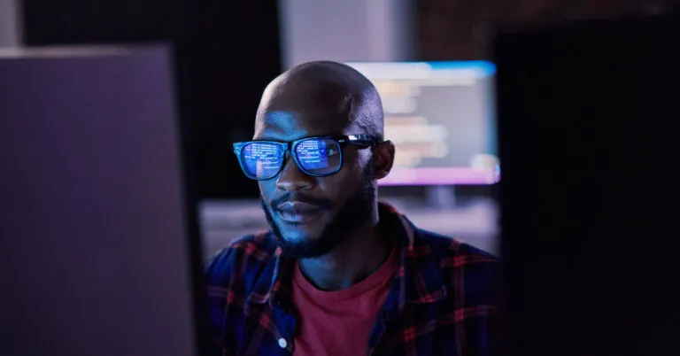Man looks at a computer monitor, and the code he is reviewing is reflected in his glasses lenses.