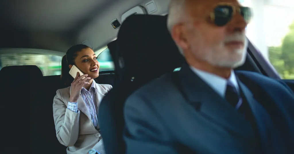 An executive rides in the back of a hired car and talks on a mobile phone with the driver visible in the front seat