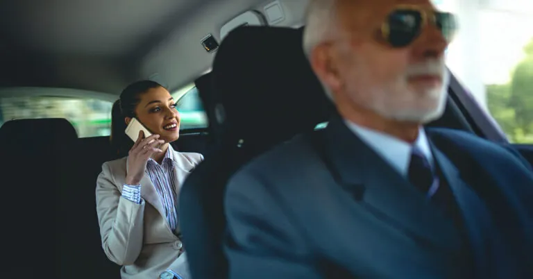 An executive rides in the back of a hired car and talks on a mobile phone with the driver visible in the front seat