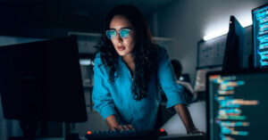 Woman stands at a desk, typing on a keyboard and looking at a monitor, with its reflection in her glasses