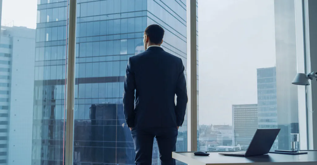A man in a suit stands, looking out of a high-rise building window with his back to the camera.