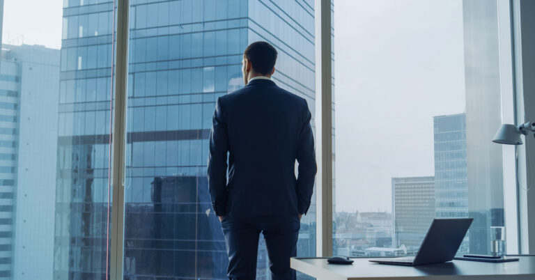A man in a suit stands, looking out of a high-rise building window with his back to the camera.