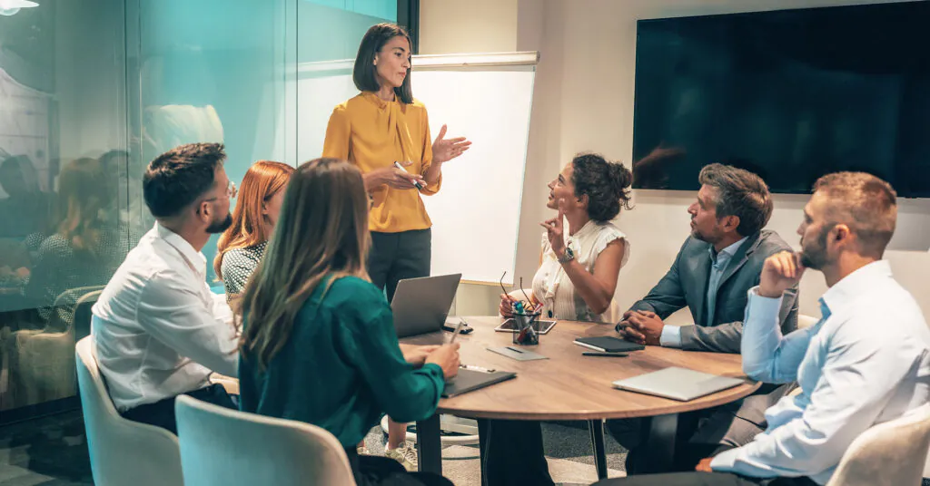 Six coworkers sit around a conference table looking up at a woman talking in front of a white board.