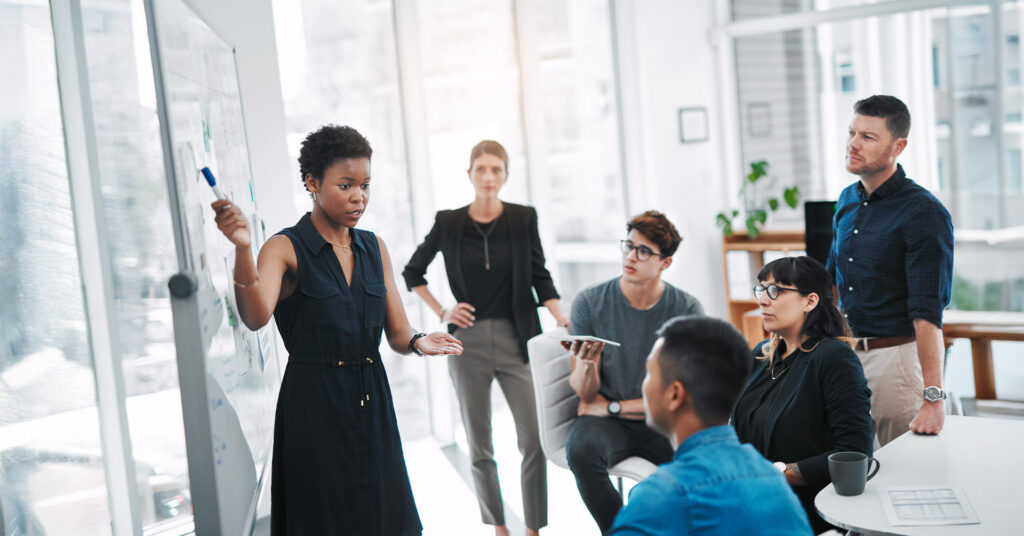 Six people are gathered in a conference room, with one woman standing in front of a white board, talking and pointing toward the board