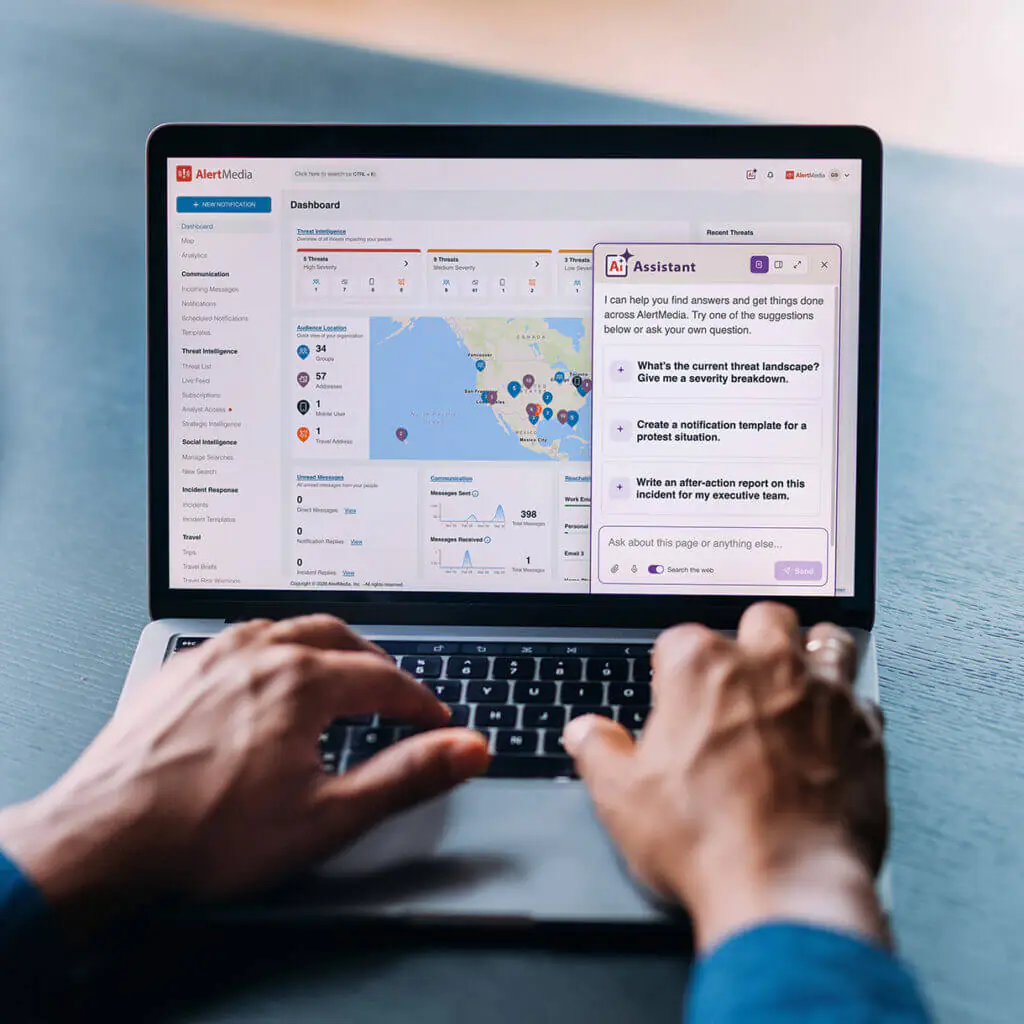 Hands type on a laptop displaying an AlertMedia dashboard with maps, incident data, and an AI assistant, conveying focused workplace safety planning.